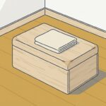 明るい室内のすみ。床にふたを閉じた木の道具箱が1つ置かれ、天面にたたんだ生成りの布が1枚のっている。