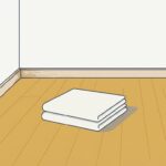 家具の何もない明るい室内の角。木の床の中央に、たたんだ生成りの厚手の布が2枚重ねて置かれている。