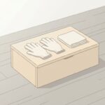 明るい室内の床に置かれた木の低い箱。天面に生成り色の布手袋が1組と、たたんだ無地の布が1枚置かれている。